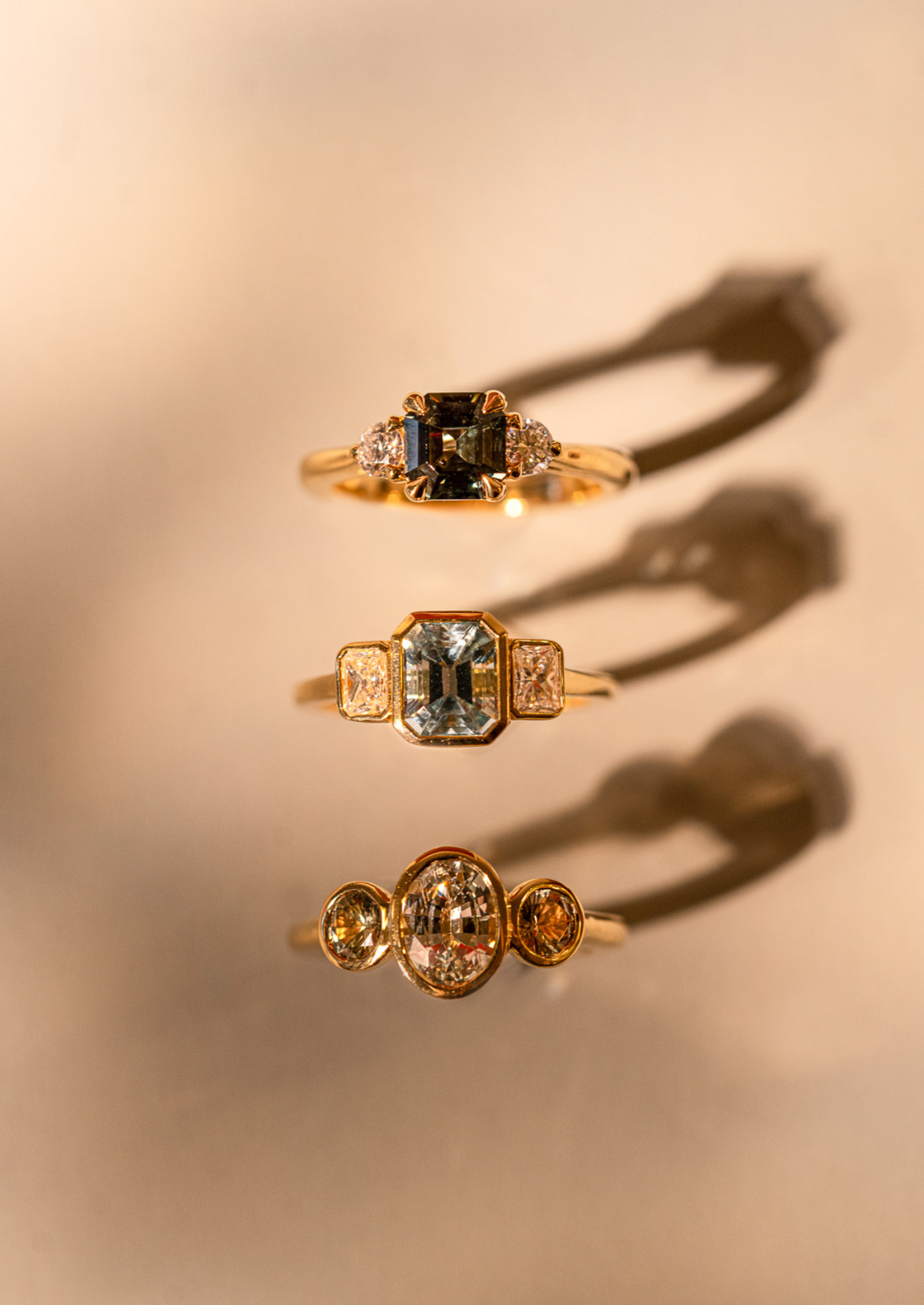 Three gold rings with differently shaped centre stones and side stones, casting long shadows.